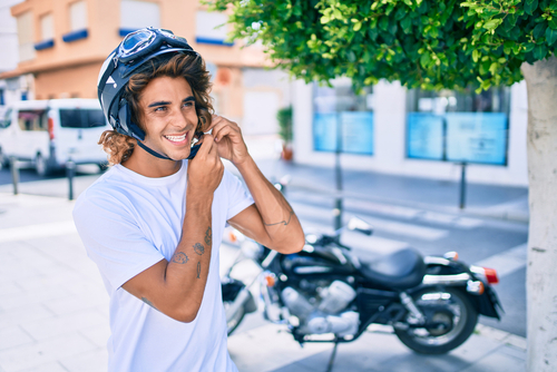Hombre joven poniendose el casco mientras esta parado junto a su moto sonriendo