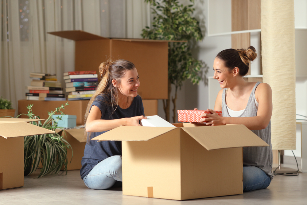 Two happy roommates talking unboxing belongings sitting on the floor